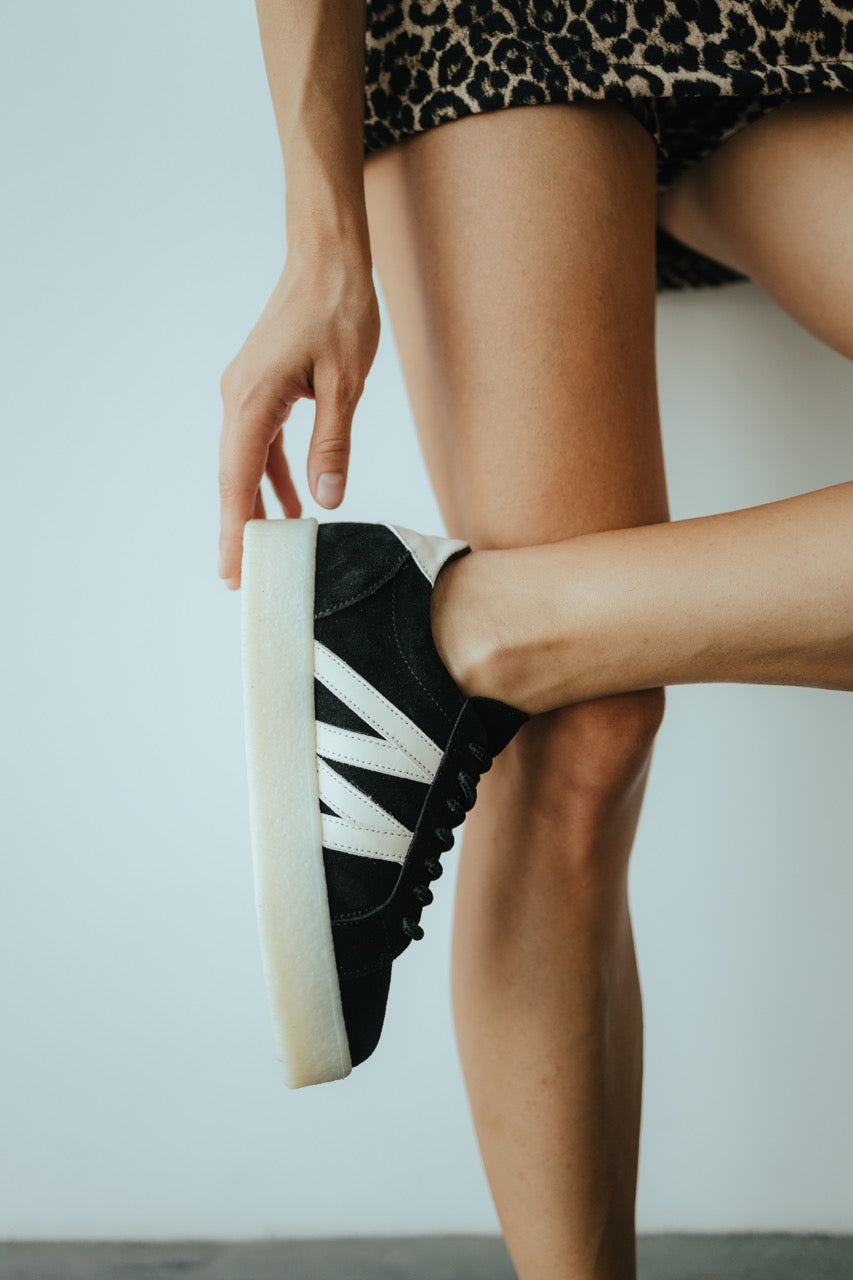 May include: Black suede trainers with white detailing. The shoes feature white soles and white, V-shaped designs on the sides. The laces are black. The trainers are on a grey surface.