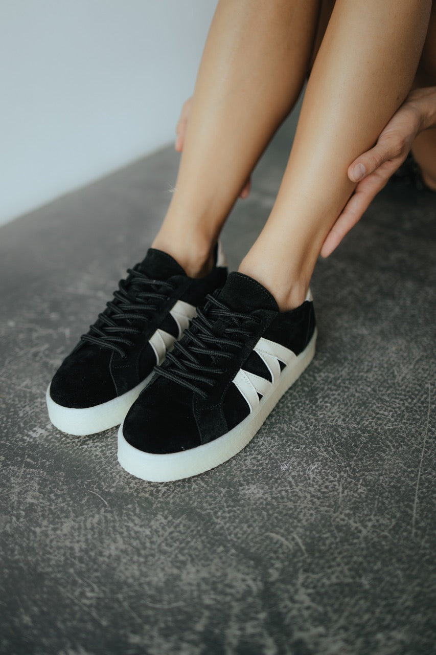 May include: Black trainers with white detailing and a thick white sole. The shoes have a stylized white "M" design on the sides. The image shows the trainers on a grey surface.